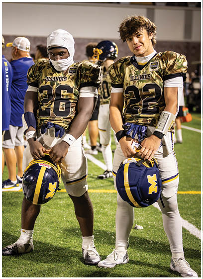 Two football players wearing camouflage uniforms and helmets pose for a picture on the field. AI generated content