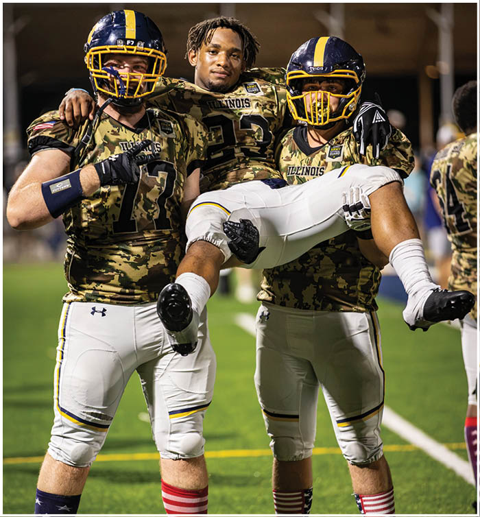 Three football players wearing camouflage uniforms are standing on a field. One player is holding another player on his shoulders, while the third player is standing nearby. AI generated content