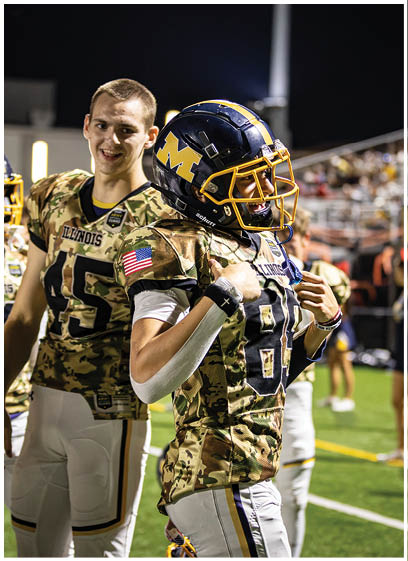 Two football players wearing camouflage uniforms are hugging each other on the field. AI generated content