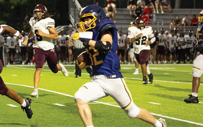 A person in a blue and yellow football uniform is running with the ball while two people in white and maroon uniforms chase them on a lit football field at night. Description generated by AI
