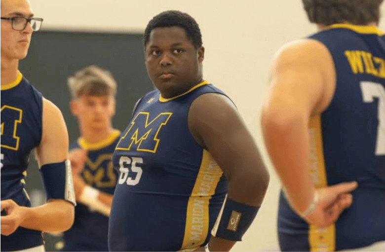 People wearing sports uniforms with the letter M and the word Wildcats are standing indoors, appearing to be in a team meeting or practice. Description generated by AI