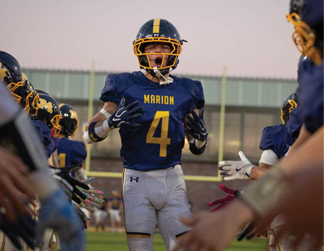 A person wearing a blue and yellow football uniform with the number 4 and the word Marion is shouting or expressing excitement while other players in similar uniforms are nearby. Description generated by AI