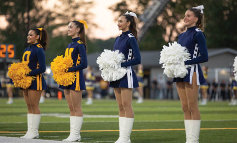 Four people wearing white boots stand in a line on a sports field near the sideline. Description generated by AI