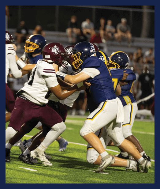 Two people in blue and white football uniforms are blocking two opponents in maroon and white uniforms during a game on a green field. Description generated by AI