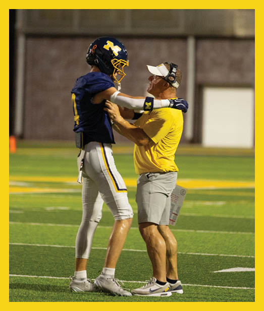 A person in a yellow shirt and visor is holding and looking up at a person in a blue football uniform and helmet on a sports field. Description generated by AI