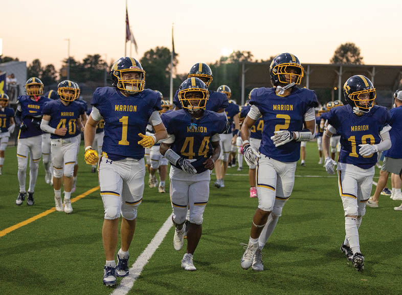Four people in blue and yellow football uniforms with helmets are running together on a green field during a game or practice. Description generated by AI