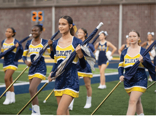 Adults dressed in blue and yellow sequined uniforms holding flags are performing on a sports field. Description generated by AI