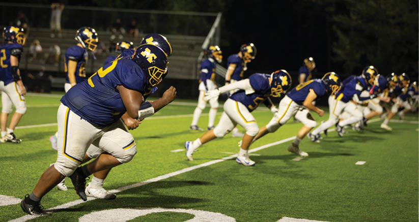 A group of people wearing blue and yellow football uniforms are lined up and starting to sprint on a green field at night. Description generated by AI