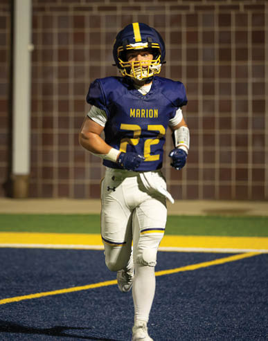A person wearing a blue and yellow football uniform with the word Marion and the number 22 on the jersey is standing on a field with a blurred background. Description generated by AI