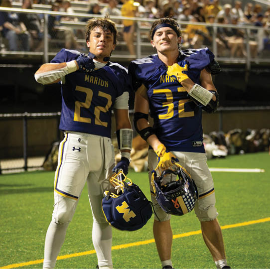 Two adults wearing blue football uniforms and shoulder pads pose together on a field with a blurred crowd in the background. Description generated by AI