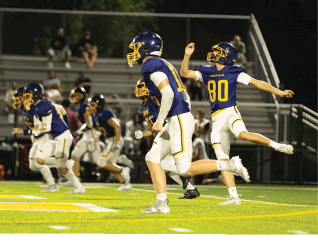 Several people in blue and white football uniforms are running on a brightly lit field with empty bleachers in the background. Description generated by AI