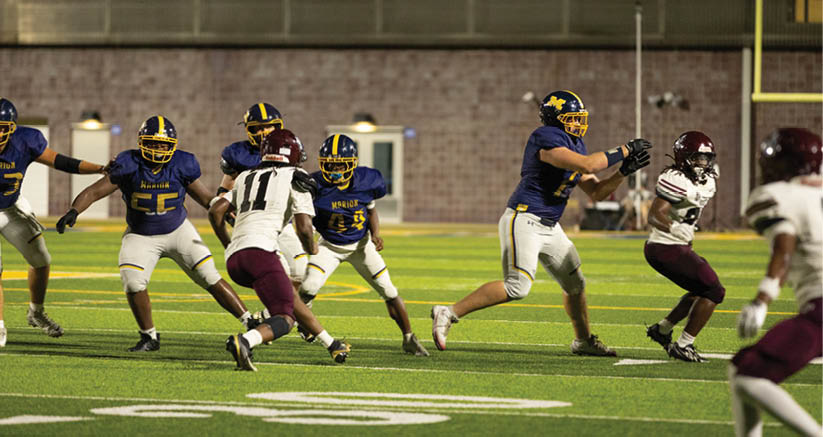 People wearing football uniforms are actively playing on a well-lit field during a game. Description generated by AI