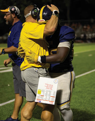 Two people, one in a yellow shirt and gray shorts and the other in a blue football uniform, are embracing on a football field at night. Description generated by AI