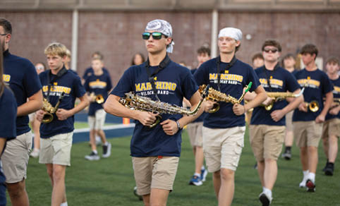 A group of people wearing matching navy blue shirts and light-colored shorts are playing saxophones while walking on a sports field. Description generated by AI