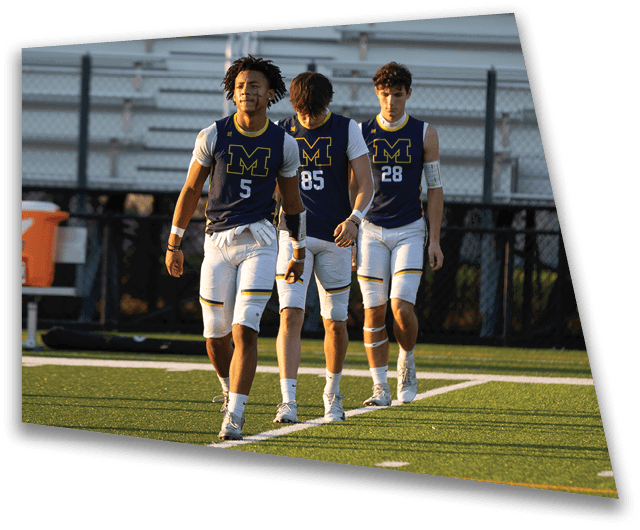 Three people wearing matching sports uniforms with the letter M and numbers walk together on a field with bleachers and water coolers in the background. Description generated by AI