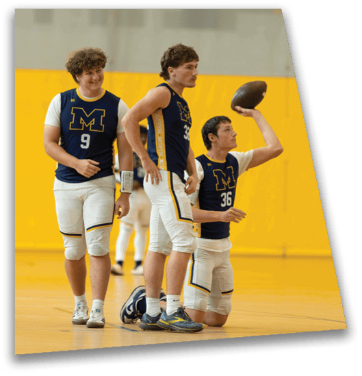 Three people wearing matching sports uniforms with the letter M are on an indoor court, with one kneeling and preparing to throw a football while the other two stand nearby. Description generated by AI