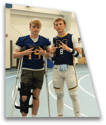 Two adults wearing football uniforms pose together on an indoor track, with one using crutches and a leg brace. Description generated by AI