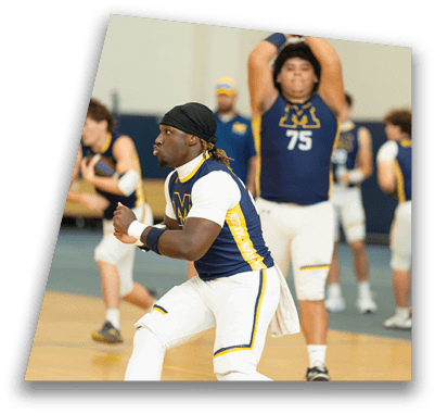 People wearing athletic uniforms with the letter M are practicing or warming up indoors, with one person in the foreground raising their arms. Description generated by AI