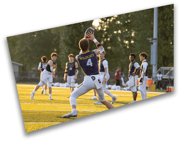 A person wearing a Wildcats jersey with the number 4 is catching a football on a sunlit field while several other players in sports uniforms watch nearby. Description generated by AI