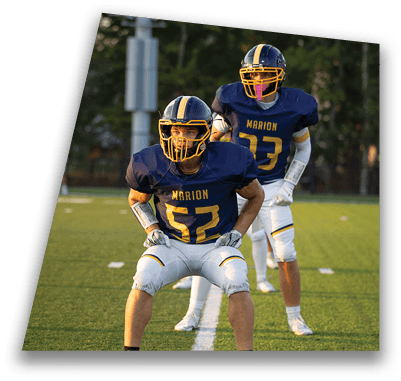 Two people wearing navy blue and yellow football uniforms with helmets are on a green field, with one person crouching in the foreground and the other standing behind. Description generated by AI