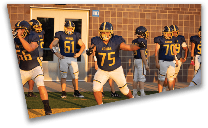 Several people wearing blue and yellow football uniforms with helmets are standing and preparing on a field near a building during sunset. Description generated by AI