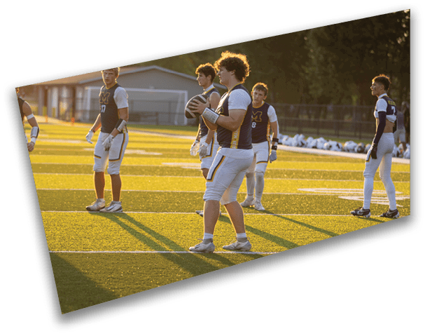 People wearing sports uniforms stand on a sunlit football field with one person holding a football, preparing for a play. Description generated by AI