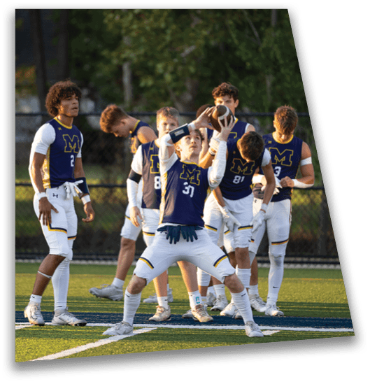 A group of people wearing matching sports uniforms are on a field with one person catching a football while others watch. Description generated by AI
