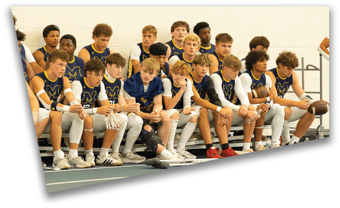 A group of people wearing matching sports uniforms sit closely together on a bench indoors, some holding footballs and one wearing a knee brace. Description generated by AI