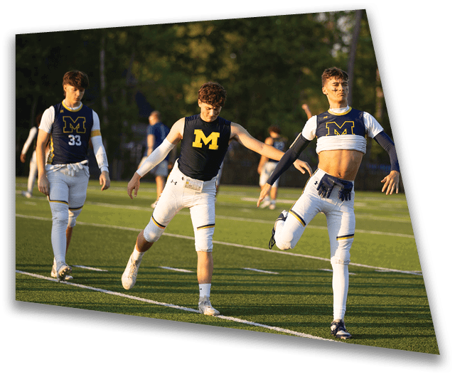 Two people wearing athletic gear with a large “M" on their shirts are stretching on a grassy sports field during daylight. Description generated by AI