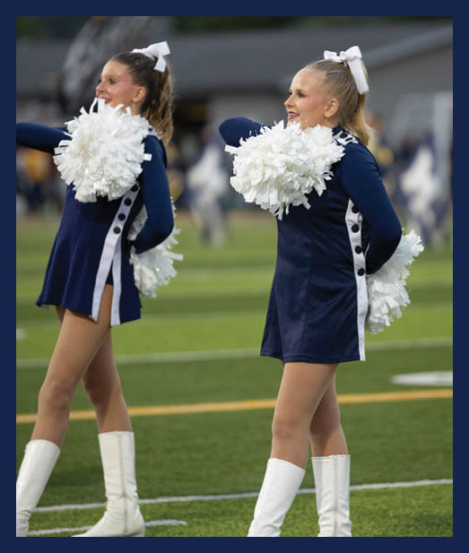 Two adults in matching navy blue cheerleading uniforms with white boots and bows perform with white pom-poms on a sports field. Description generated by AI