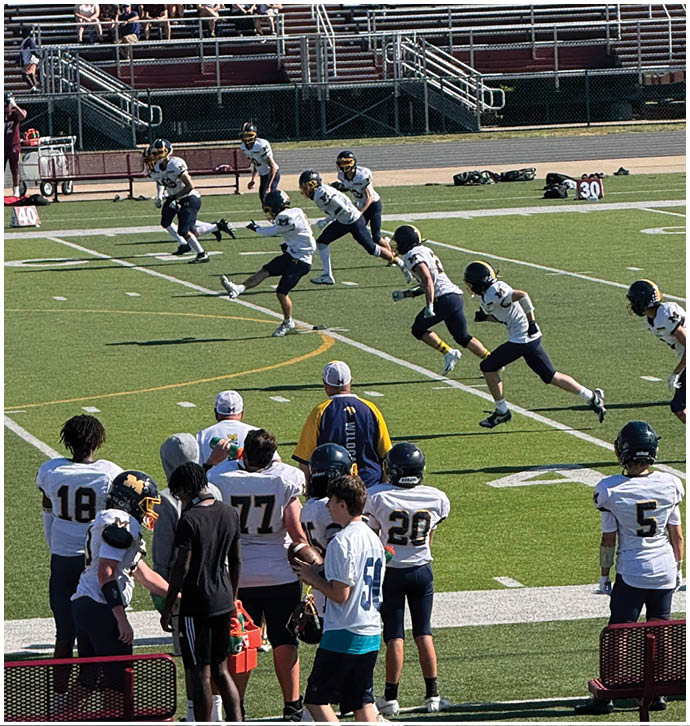 Players in football uniforms are running on a field while others watch from the sidelines and spectators sit in the bleachers. Description generated by AI
