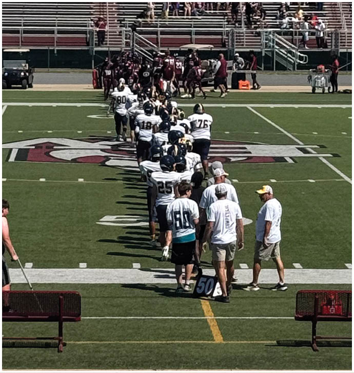 People in sports uniforms and casual clothing are lined up on a football field near the 50-yard line with empty bleachers and a sign reading “HOME OF THE MAROONS" in the background. Description generated by AI