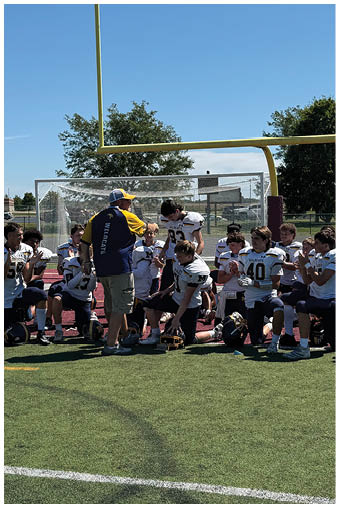 A group of children in football uniforms are gathered on a field with a coach in a cap and sportswear giving instructions near a goalpost. Description generated by AI