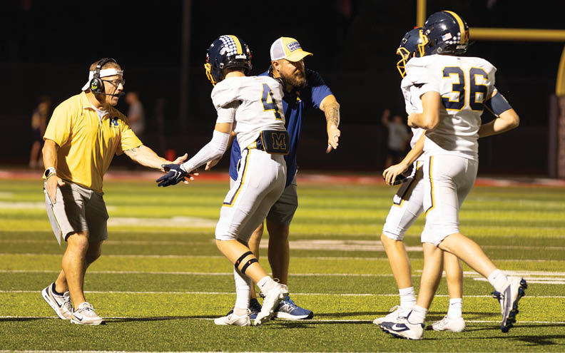 A coach wearing a cap and blue shirt is instructing three football players in white uniforms and helmets on a field at night. Description generated by AI