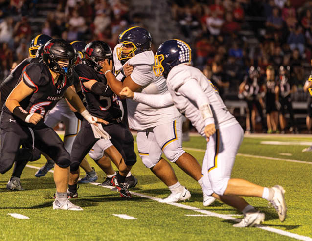 People in football uniforms are engaged in a play on a well-lit field with a crowd in the background. Description generated by AI