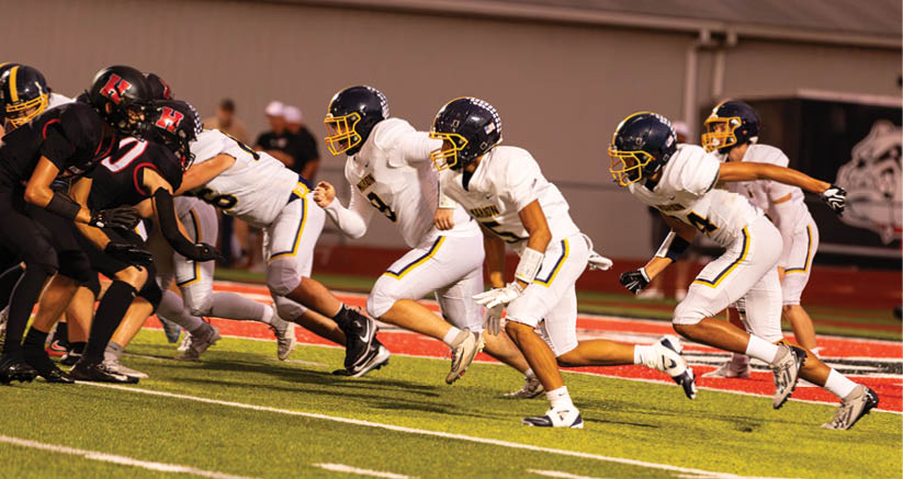 Several people in white football uniforms and helmets are running on a field toward a group of opponents in black uniforms during a game. Description generated by AI