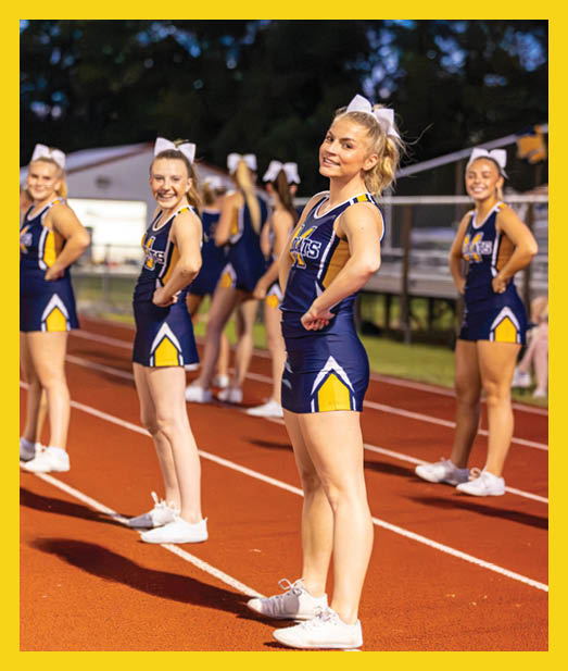 Two people wearing cheerleading uniforms with large white bows in their hair are smiling outdoors at night with others in similar attire blurred in the background. Description generated by AI