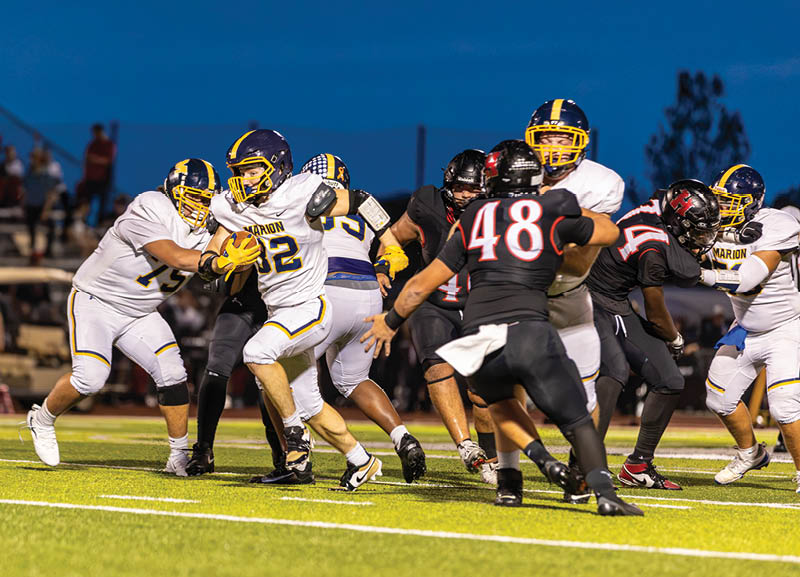 Players in football uniforms are actively engaged in a game on a well-lit field during dusk with spectators in the background. Description generated by AI