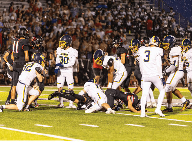Players in white and black football uniforms are engaged in a play on a brightly lit field with a crowd in the background. Description generated by AI