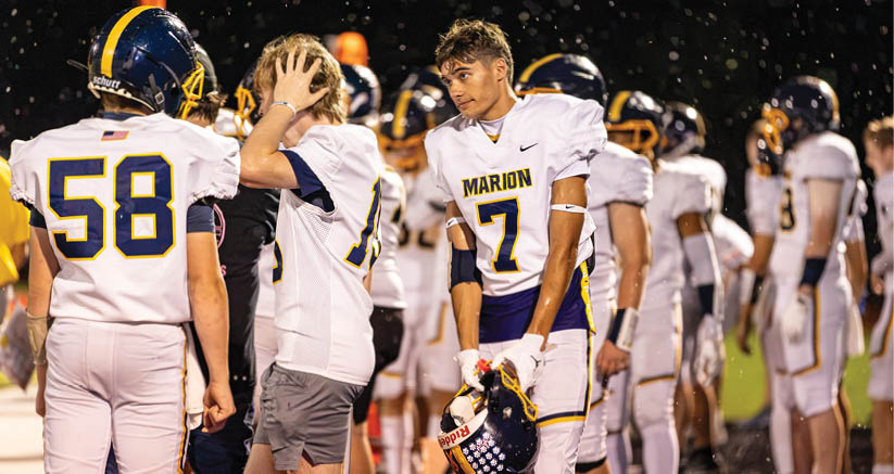 People in football uniforms stand on a field, with one person holding a helmet and others appearing to prepare or rest during a game or practice. Description generated by AI