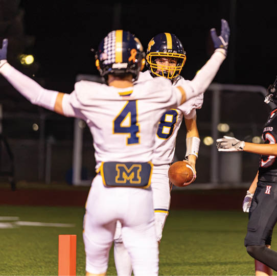 Two people in white football uniforms celebrate while a person in a black football uniform stands nearby on a field at night. Description generated by AI