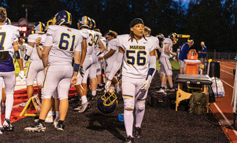 A group of people in football uniforms are gathered on a field with one person walking toward the camera holding a helmet. Description generated by AI