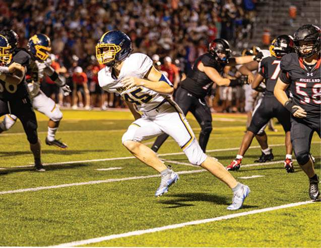 An adult in a white football uniform is running on a field while adults in black football uniforms are positioned behind. Description generated by AI
