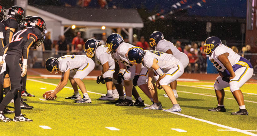 People in football uniforms are lined up on a field preparing for a play during a nighttime game. Description generated by AI
