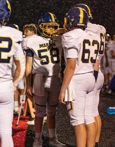 People wearing football uniforms with helmets stand together on a field at night with visible rain or snow falling. Description generated by AI