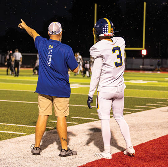 An adult wearing a blue shirt and beige shorts is standing next to a person in a white football uniform with the number three on the back on a football field at night. Description generated by AI