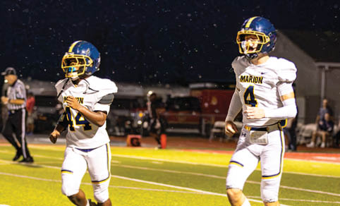 Two people wearing white football uniforms and blue helmets are running on a well-lit football field at night. Description generated by AI