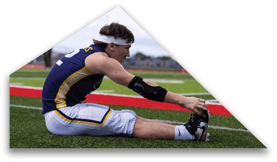 A person wearing athletic gear and a headband is sitting on a sports field stretching their leg while holding their foot. Description generated by AI