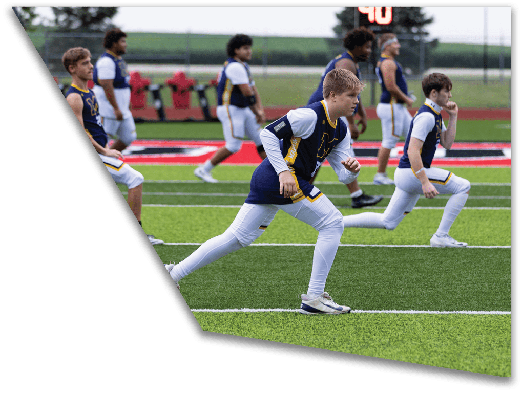 People in sports uniforms are warming up or stretching on a green artificial turf field with a scoreboard in the background. Description generated by AI