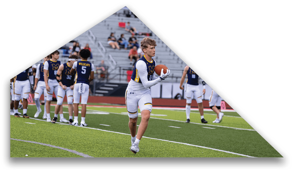 An adult wearing a white and navy sports uniform is holding a football on a green field with other players in similar uniforms standing in the background. Description generated by AI